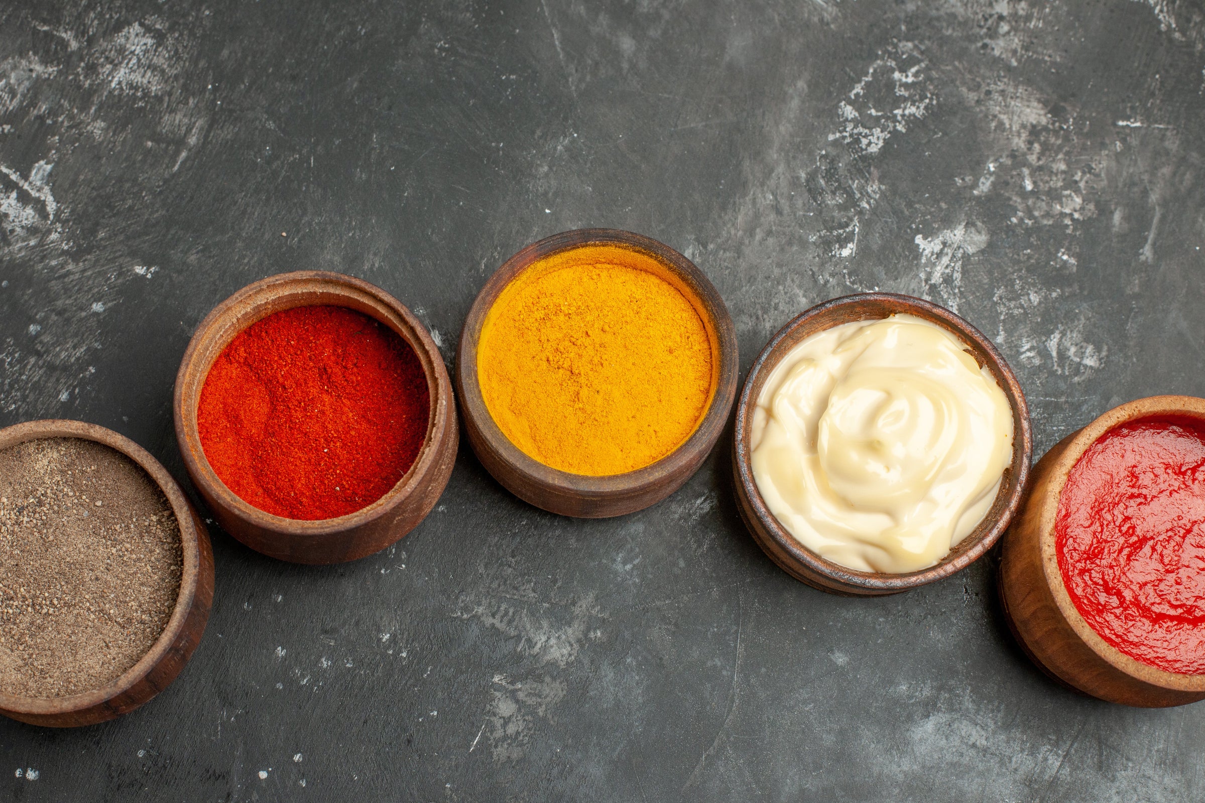 Quatre petits pots en bois contenant différentes sauces et épices, alignés sur une surface sombre texturée. Du rouge vif au jaune curry, en passant par un blanc crémeux, chaque pot offre une couleur et une texture unique.